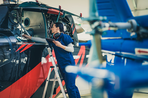 Serious female mechanic in uniform standing on ladder near jet and holding tablet, side view. Aircraft engineer working in helicopter hangar, medium shot