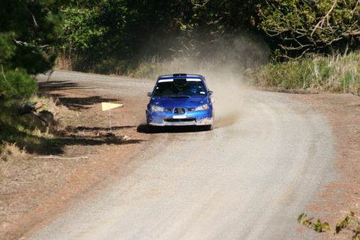 Blue rally car racing on a gravel road, gravel and dust spraying out as it comes round the corner at speed.