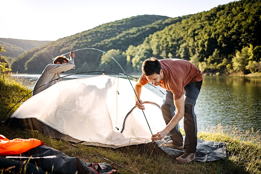 Two men on a camping trip adjusting a tent while standing and crouching by the lake in nature
