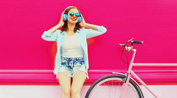 Summer portrait of happy smiling young woman listening to music in headphones with bicycle on pink background