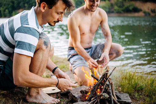 Two men are preparing sausages on a campfire by the lake