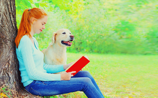 Young woman reading book with Golden Retriever dog sitting near tree in summer park