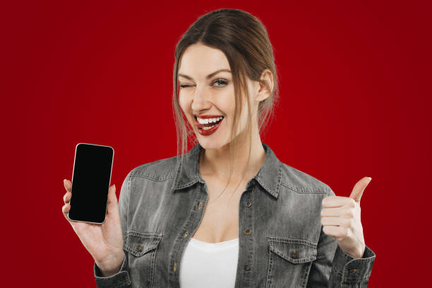 istock Satisfied female phone user on red background winking raising thumb up showing blank screen mockup 1403828278
