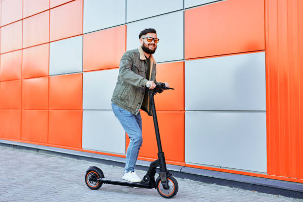 istock Bearded man in sunglasses posing on electric scooter over orange wall background. Man riding kick scooter. 1403828443