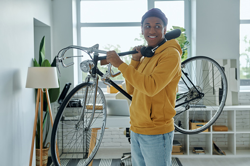 Cheerful young multiracial man carrying bicycle on shoulder while standing in office