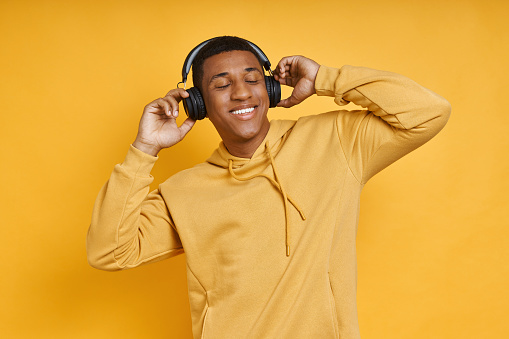 Cheerful mixed race man in headphones enjoying music while standing against yellow background
