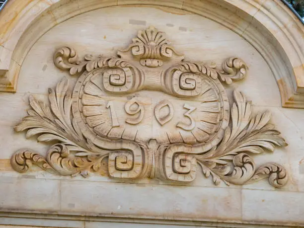 The year 1903 is engraved on an architectural feature on the facade of an old building. The condition is very good. The building is very old and the exterior is renovated.
