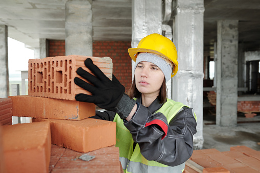 Young serious female builder in protective helmet and workear taking brick from top of huge stack prepared for building work