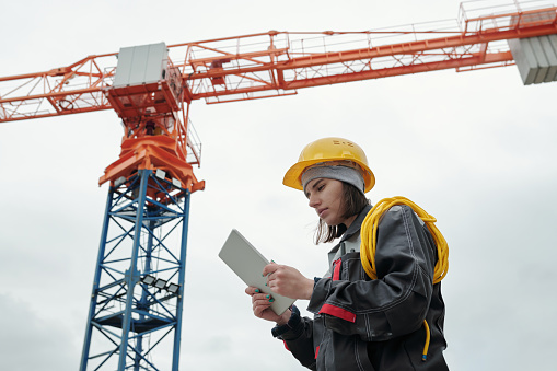 Young serious female engineer in workwear and protective helmet looking at screen of tablet while standing against working crane