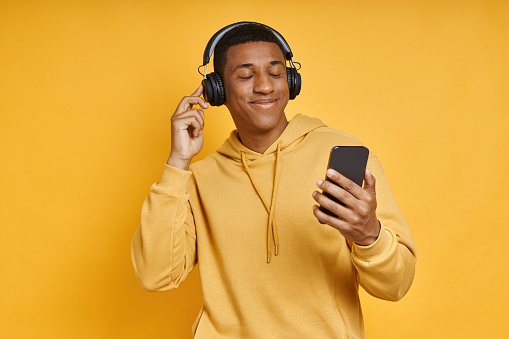 Cheerful mixed race man with headphones using smart phone while standing against yellow