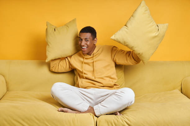 istock Happy mixed race man holding pillows while sitting on the couch with yellow background 1403847372