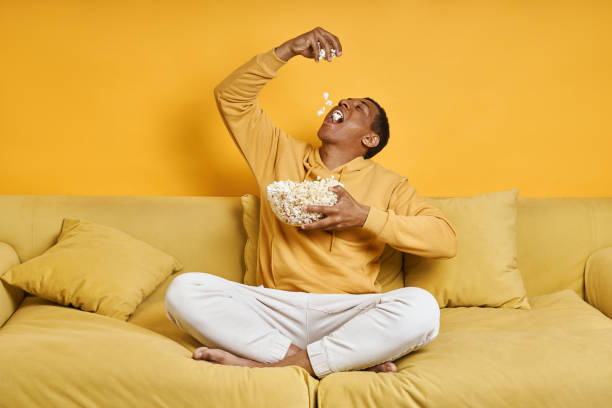 Playful mixed race man enjoying popcorn while relaxing on the couch with yellow background stock photo