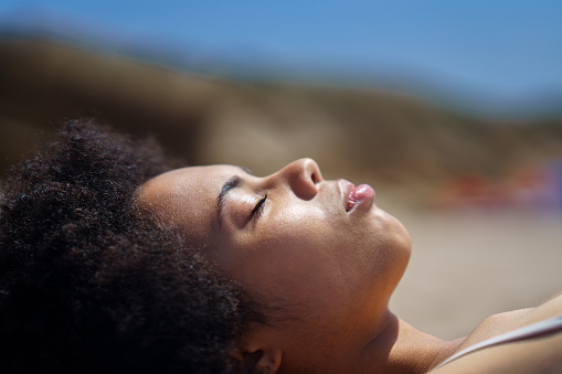 Side view of peaceful black female tourist with Afro dark hair, relaxing on sandy beach with closed eyes, and sunbathing on sunny day during summer holiday
