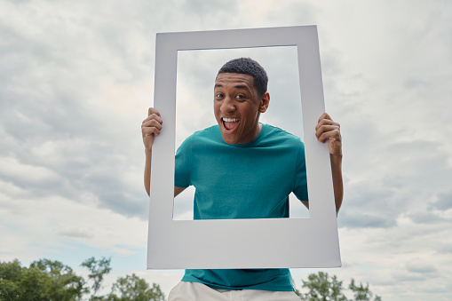 Cheerful mixed race man looking through a picture frame while standing outdoors