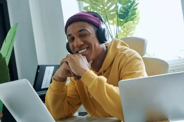 Cheerful mixed race man in headphones sitting at his working place and winking