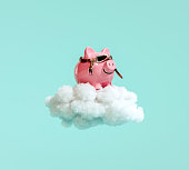 Piggy bank with pilot glasses flying on white fluffy cloud on turquoise blue background