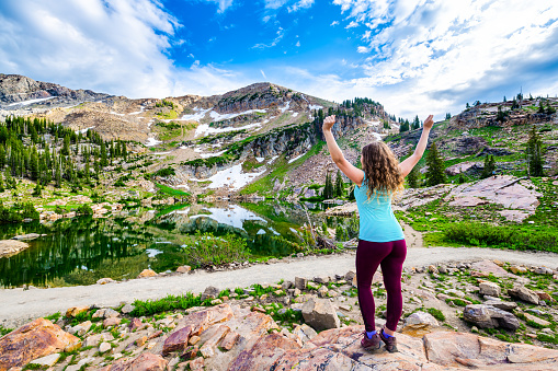 Albion Basin, Utah summer with happy woman back standing looking at view reflection of water on Cecret Lake in Wasatch mountains with rocks, snow and green color