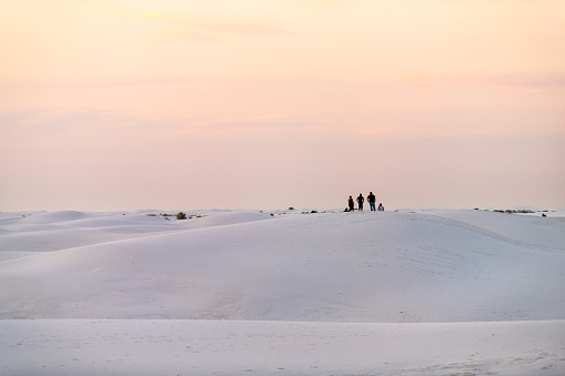 White sand dunes in New Mexico with silhouette of group of people far distant in background during pastel pink and yellow sunset sunlight in sky