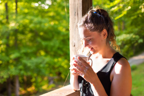 夏の朝の木造キャビンコテージ田舎の田舎のリトリートで家の素朴な木製のポーチで日の出の日差しでコーヒーや紅茶を飲んで幸せな笑顔の女性 - 16025 ストックフォトと画像