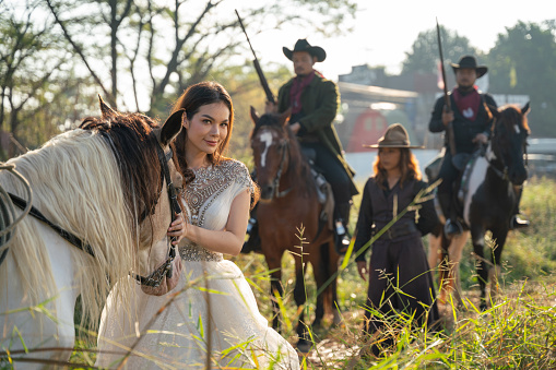 Pretty woman with white dress stand near horse and Asian man with cowboy costume stay on horse that look like he guard or protect her in field near village.