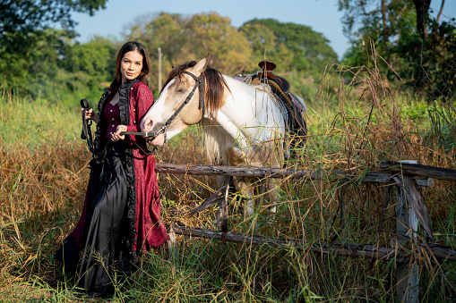 Pretty young Asian woman stand and lay to fence near white horse also look at camera in field.