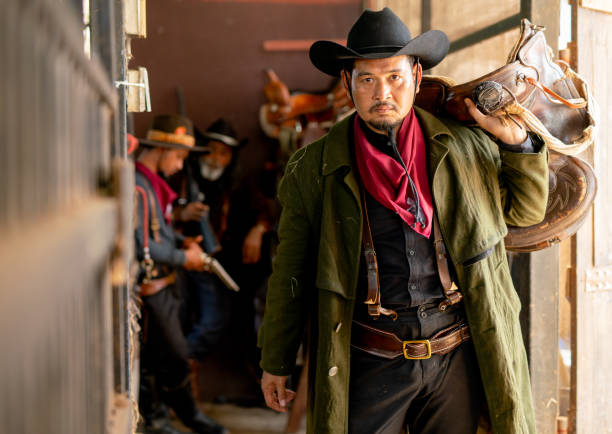 istock One cowboy hold saddle and look at camera while other two man check the gun in background of stable. 1403856984