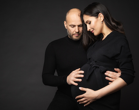 Happy Pregnant Couple looking at Belly. Mother and Father Love expecting Baby during Pregnancy. Family waiting Child Birth over Black Studio Background. Man Hands on Pregnant Woman Abdomen
