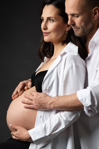 pareja embarazada con camisas blancas abrazando el vientre sobre el fondo negro. a la madre y al padre les encanta esperar un bebé durante el embarazo. familia en espera de parto. manos del hombre en la mujer embarazada abdomen desnudo - two parent family naked men couple fotografías e imágenes de stock