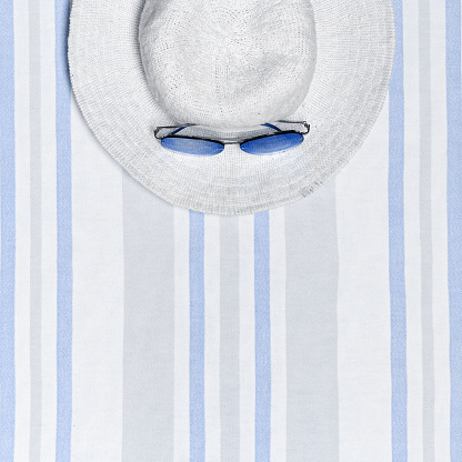 Minimal summer beach concept top view wide-brimmed sun hat and sunglasses on striped beach towel, pastel blue white background. Summertime party, travel, holiday card or invitation. Happy Summer day.