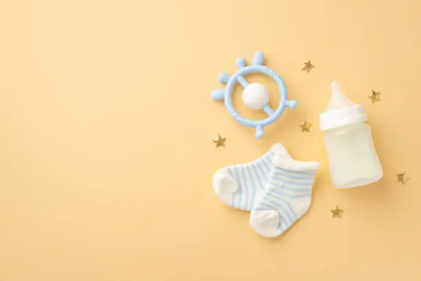 Photo of Baby accessories concept. Top view photo of blue rattle tiny socks milk bottle and gold stars on isolated pastel beige background with copyspace