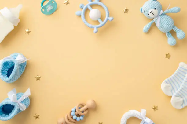 Photo of Baby concept. Top view photo of blue teddy-bear toy rattle socks milk bottle teether soother knitted shoes and gold stars on isolated pastel beige background with empty space in the middle