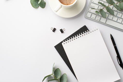 Business concept. Top view photo of workspace keyboard black and white note pads pen binder clips cup of coffee on saucer and eucalyptus branches on isolated white background with empty space