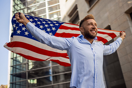 Portrait of motivated middle eastern man who holding USA flag and looking at camera. celebrating national holiday, die-hard fan with long hair and tattoo on yellow background