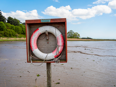 A red and white lifebelt hanging on a wooden stand beside the mudflats at low tide on the River Deben at Ramsholt, Suffolk, Eastern England.
