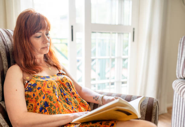 Mature woman reading a book while sitting alone on a chair in her living room at home