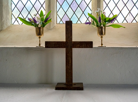 A small, plain wooden cross standing on a church altar with two small brass chalices filled with spring flowers on the window sill above. (Suffolk, Eastern England.)