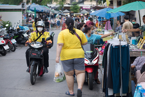 Young thai woman in yellow shirt with food in bag is walking in street wit local market in residential district in Bangkok Chatuchak. In street are market stalls and some people. An epxress delivery person is driving motorcycle. Woman is wearing casual clothing