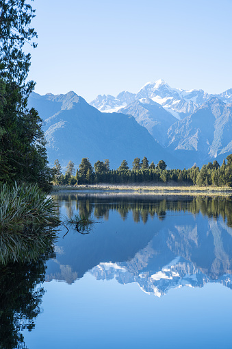 Perfect reflection in Lake Matheson surrounded by beautiful natural forest under blue sky with view to Southern Alps and Mount Cook, South Island new Zealand.