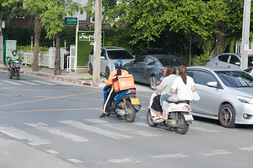 Motorcycle drivers and a delivery person are waiting for turning into a street in Bangkok Chatuchak. They are standing in center of street and let some cars passing by. Delivery service person is female and has a box in back. Service is branded Shopee Food. Other women on motorcyce are not wearing a crash helmet. In left background another express delivery person is driving