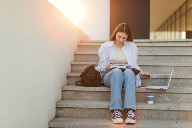 A young woman with long red hair sits on a city staircase, surrounded by brick architecture, writing in a notebook while holding a smartphone.