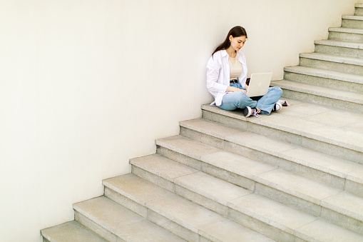 Beautiful female student is sitting on stairs in front of university building and using laptop. She is wearing white shirt and trousers and learning between classes