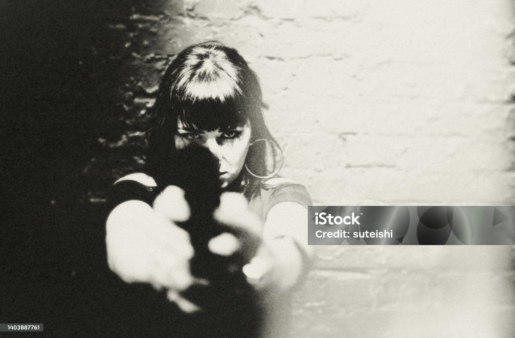It happened after 10 pm! Portrait of a young woman with a pistol. She looks directly, focused and sternly at her opposite. She is ready for anything. Dark and mysterious, Film Noir style. Target Shooting Stock Photo