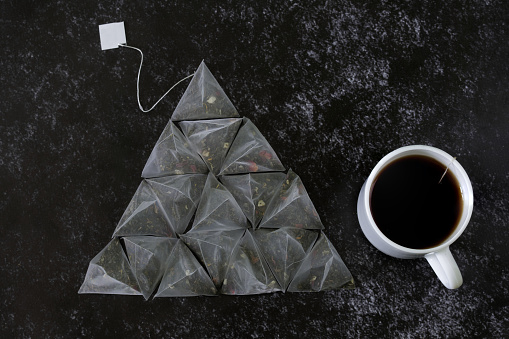 Set of pyramid tea bags with green flavored tea, with the addition of fruits, and a white mug on the dark textured surface of the kitchen table. Pyramid shape. Close-up