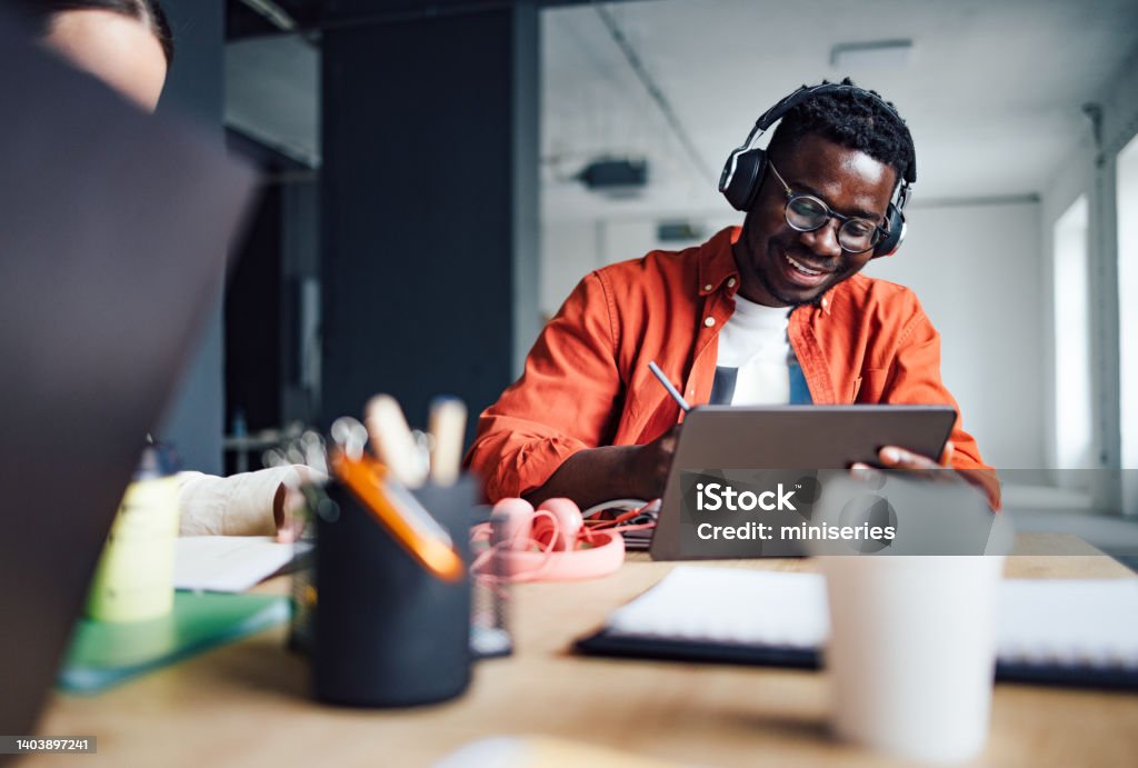 University Students Studying Together In The Library Happy African-American student sitting at desk with his unrecognizable friend while listening online lesson on his digital tablet and writing homework in a notebook. E-Learning Stock Photo