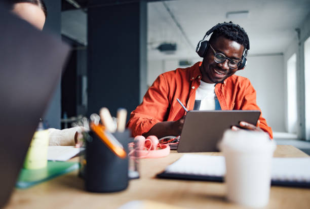 University Students Studying Together In The Library Happy African-American student sitting at desk with his unrecognizable friend while listening online lesson on his digital tablet and writing homework in a notebook. education building stock pictures, royalty-free photos & images