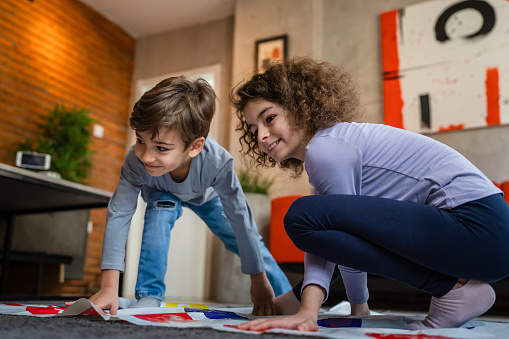 Brother and sister siblings small caucasian boy and girl child play twister game on the floor at home alone real people family growing up leisure concept copy space