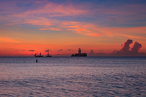 The sun sets off the West Coast of Aruba in the Caribbean Sea. It turns the horizon into gold. A couple of sailboats and an oil drilling rig can be seen on the horizon. Photo shot at sunset.