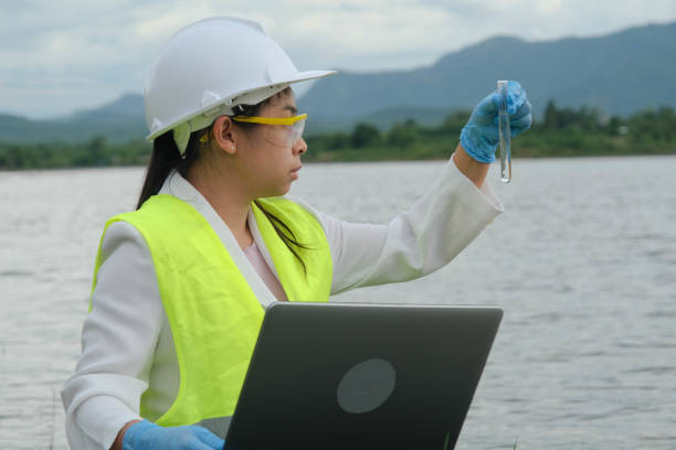 手袋を着用した女性の環境保護主義者は、川の水のサンプルを採取して天然水域の汚染物質を調べ、ラップトップにデータを記録します。水とエコロジーの概念。 - 環境保護主義者 ストックフォトと画像