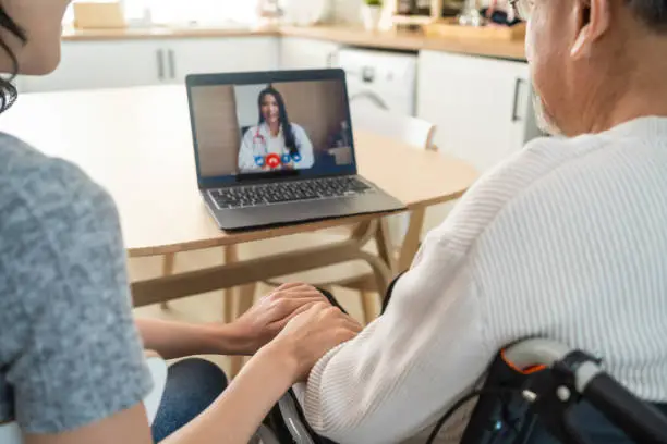 Photo of Asian senior male and daughter video call with doctor in living room. Elderly old mature grandfather sitting on table with young granddaughter, using computer laptop consult with general practitioner.