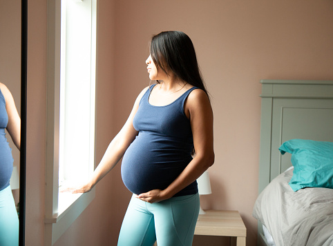 Young very pregnant latina looking out the window in bedroom, relaxed, touching her baby bump.